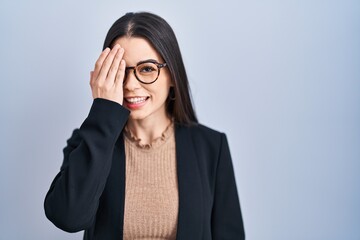 Young brunette woman standing over blue background covering one eye with hand, confident smile on face and surprise emotion.