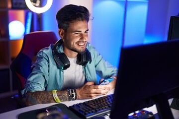Young hispanic man streamer using computer and smartphone at gaming room