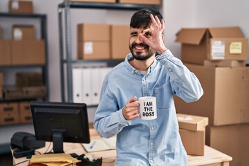 Young hispanic man with beard working at small business ecommerce drinking from boss cup smiling happy doing ok sign with hand on eye looking through fingers