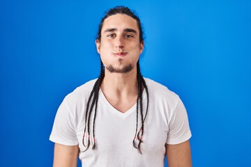 Hispanic man with long hair standing over blue background puffing cheeks with funny face. mouth...