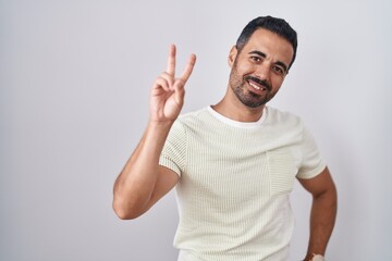 Hispanic man with beard standing over isolated background smiling looking to the camera showing fingers doing victory sign. number two.