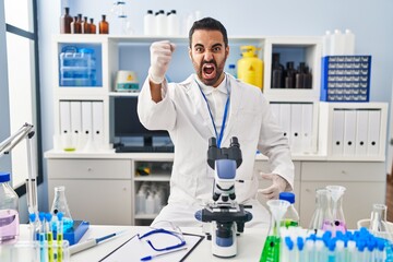 Young hispanic man with beard working at scientist laboratory angry and mad raising fist frustrated and furious while shouting with anger. rage and aggressive concept.