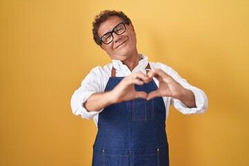 Middle age hispanic man wearing professional cook apron smiling in love doing heart symbol shape...