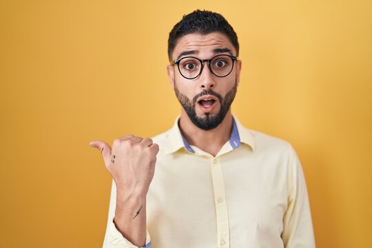 Hispanic young man wearing business clothes and glasses surprised pointing with hand finger to the side, open mouth amazed expression.