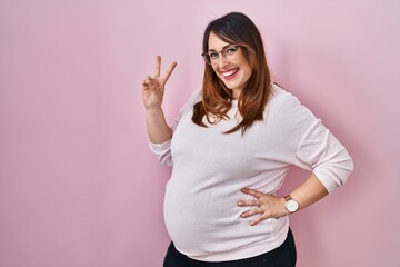 Pregnant woman standing over pink background smiling looking to the camera showing fingers doing...
