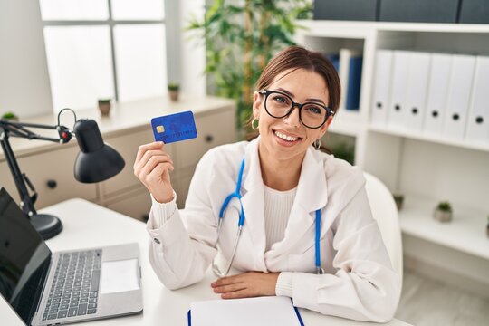 Young hispanic woman wearing doctor uniform holding credit card at clinic - Powered by Adobe