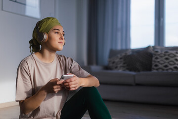 Young woman with cancer siting at home and scrolling her phone and listening the music.