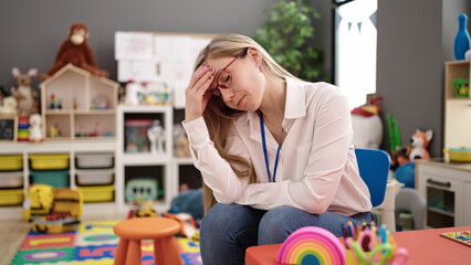Young blonde woman preschool teacher standing with relaxed expression at kindergarten