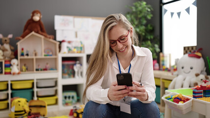 Young blonde woman preschool teacher smiling confident using smartphone at kindergarten