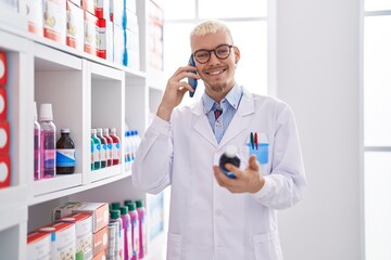 Young caucasian man pharmacist talking on smartphone holding medicine bottle at pharmacy