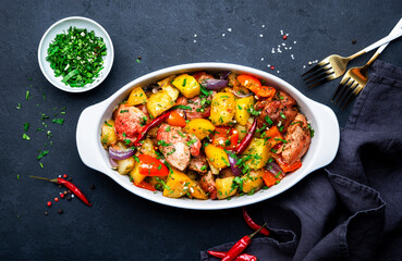 Baked turkey drumsticks fillet with potatoes, paprika, onions, herbs and spices in baking dish, black table background, top view