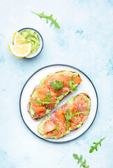 Avocado salmon sandwich or toast on rye bread with guacamole sauce, young arugula and sesame seeds on plate, blue table background, top view