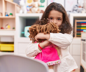 Fototapeta na wymiar Adorable hispanic girl hugging doll standing at kindergarten