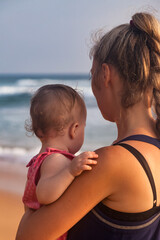 Rear view mom with baby in arms standing on tropical beach at sea background. Female with little child in summer travel vacation. Concept of motherhood and recreation with children. Copy text space