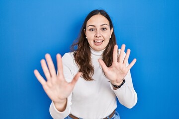 Young hispanic woman standing over blue background afraid and terrified with fear expression stop gesture with hands, shouting in shock. panic concept.