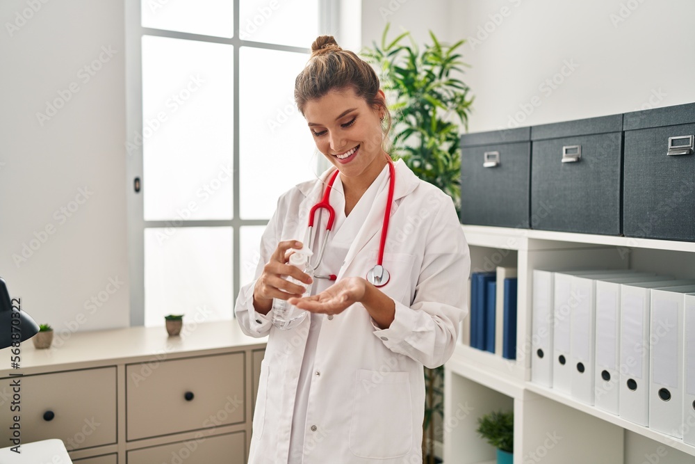 Poster Young blonde woman wearing doctor uniform using sanitizer gel hands at clinic