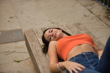 Young beautiful woman with straight brown hair, orange top and jeans, lying on a wooden bench, smiling happily. Concept fashion, beauty, trend, happiness, relax, millennial.