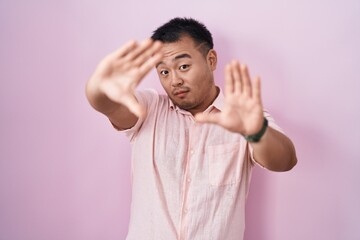 Chinese young man standing over pink background doing frame using hands palms and fingers, camera perspective