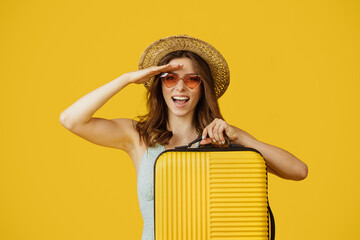Young woman traveller with straw hat carrying suitcase and looking at camera, standing over yellow...