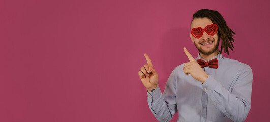 Concept of Valentine's Day. Young Caucasian man with dreadlocks isolated on pink background. Guy wears heart-shaped glasses and bow tie. Points with both hands to side and smiles. Web banner.