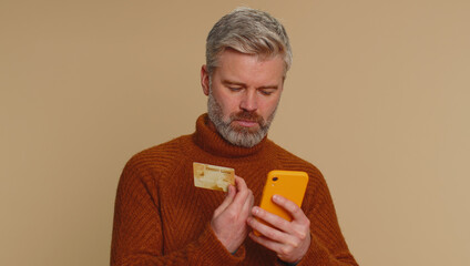 Sincere middle-aged man customer using credit bank card and smartphone while transferring money, purchases online shopping, payment. Finance and internet. Senior guy isolated alone on beige background