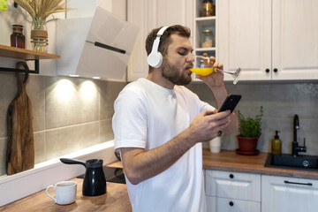 Male athlete in headphones drinking orange juice in morning and reading news on internet using his mobile phone.