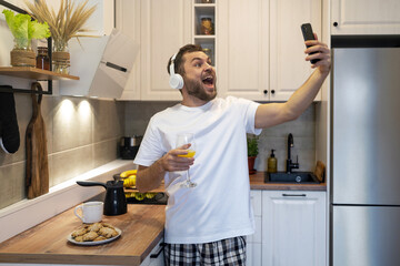 Man in headphones standing in kitchen in morning taking selfie using his phone and having fun.