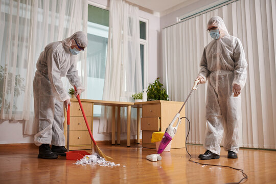 Cleaners in protective suits wiping and vacuum cleaning floor in house of client