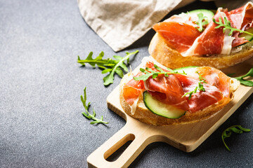 Appetizer crostini, open sandwiches with cream cheese, prosciutto, cucumber and arugula at cutting board.