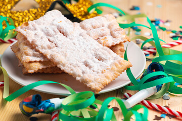 Close up of a plate of Chiacchiere over a wooden table with carnival trumpet, carnival mask, confetti, streamers and candy canes