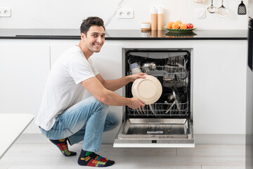 Young smiling man putting dirty plates in dishwasher machine in the kitchen. Household and...