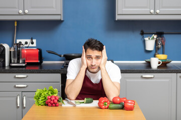 Tired and stressed man in red apron sits at the table and tries to cook, but it is too complicated...