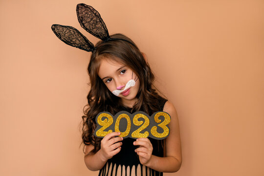 The girl holds in her hands the golden numbers of the new year 2023. A child dressed up as a black rabbit on a beige studio background.