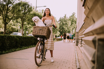 A young woman with a bichon dog in a bicycle basket takes a leisurely ride