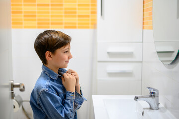 Boy puts on the collar of his denim shirt in front of the toilet mirror