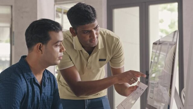 Medium shot of two Indian UX designers discussing mobile app wireframe sketch on paper and computer screen while working on project together in IT company office