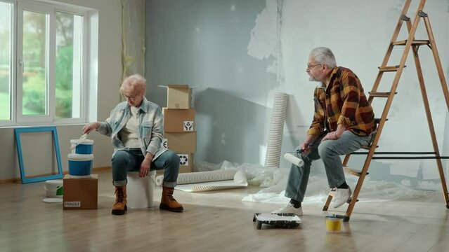 Tired elderly man is calling a master to repair an apartment using a mobile phone. Couple of aged is rejoicing at the solved problem and the help of the master. Ladder, cardboard boxes and window in