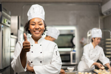 Selective focus of an African female baker in a chef uniforms, standing with arms folded while thumbs up and smiling at the camera, with blurred colleagues in the background. Copy space on right side.