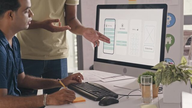 Indian UX designer discussing mobile app wireframe on computer screen with colleague while working together on project in IT company office