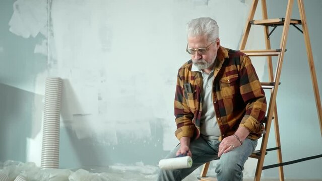 Tired elderly man is sitting on a ladder and calling a master to repair an apartment using a mobile phone. Aged pensioner is rejoicing at idea and solved problem with help of the master.