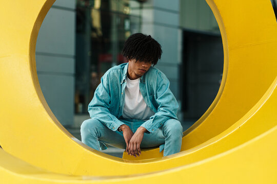 joven adolescente sentado en una estructura amarilla circular en la ciudad, al atardecer, mirando a la cámara con afro y ropa urbana.
