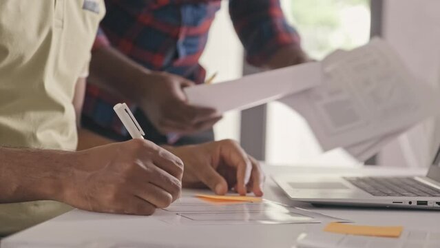 Cropped shot of two mobile app developers discussing UX project sketches on paper and laptop screen and taking notes while cooperation during workday in office