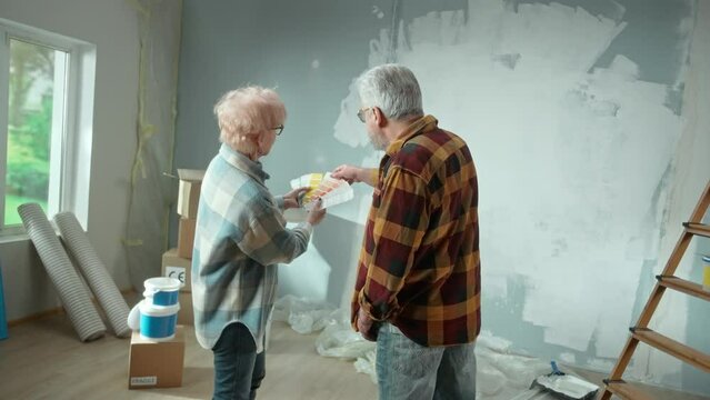 Elderly man and woman are choosing paint color on color palette. Aged couple is discussing color of wall painting, during renovation of apartment, rear view. Concept of repair, decoration, interior
