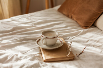 Fototapeta na wymiar A cup of hot cappuccino, a book, glasses on the bed. Breakfast. Cozy house. Decor, details.