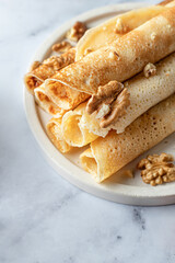 Rolled crepes or thin pancakes with honey and nuts on a white marble background close up, text space. Maslenitsa or Pancake day traditional food concept