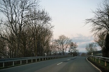 Landstraße zwischen Bäumen hinter Leitplanken vor Himmel bei Abendrot im Winter