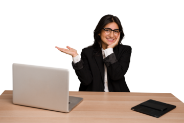 Young indian woman in a table with a laptop and tablet isolated holds copy space on a palm, keep hand over cheek. Amazed and delighted.