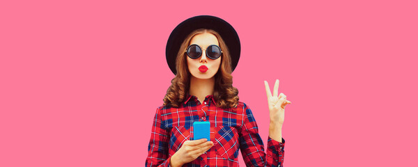 Portrait of young woman blowing her lips with phone wearing black hat and red shirt on blue background
