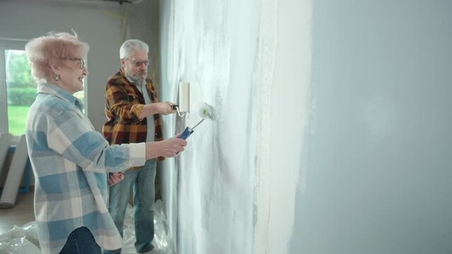 Elderly man and woman are painting wall with white paint using paint rollers. Couple of pensioners is making repairs in their apartment against backdrop of window with bright sunlight and have fun
