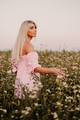Fototapeta na wymiar Vertical photo blonde woman posing in the big endless field of daisies in summer evening. Lady wear pink dressed, looking aside smiling, touching the plant green blossoming flower. Moody atmosphere 